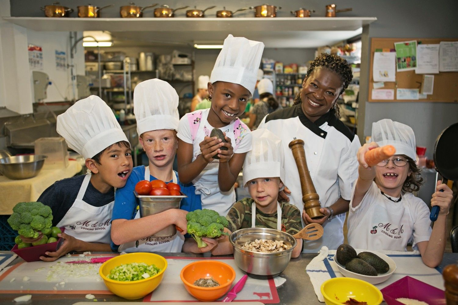 A chef and a group of children in chef hats holding fresh vegetables in a teaching kitchen